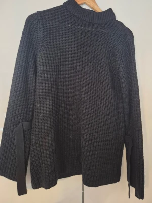 Svart stickad tröja med knytband - Säljer en svart stickad tröja med grov ribbad struktur och högt krage. Tröjan har långa ärmar och coola knytband vid ärmsluten som ger en extra detalj. Perfekt för dig som gillar en enkel men ändå unik look.