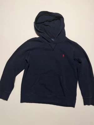 Mörkblå Polo Ralph Lauren Hoodie. - Mörkblå Polo Ralph Lauren Hoodie med huva och känguruficka framtill. Liten röd broderad logga på bröstet. Tillverkad i mjukt bomull. 