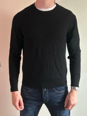 Svart stickad tröja från ASKET - Klassisk svart stickad tröja från ASKET med rund halsringning och ribbade muddar. Tröjan har en enkel och stilren design som passar till det mesta. Perfekt basplagg för garderoben och lätt att matcha med jeans eller byxor.