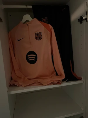 Ljusrosa långärmad Nike topp med matchande blå byxa  - Säljer en ljusrosa långärmad topp från Nike med FC Barcelona-logga och Spotify-tryck på bröstet. Tröjan har half zip och är perfekt för dig som gillar sportig stil. Passar både till träning och chill.