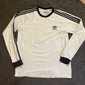  Adidas Originals 3-Stripes Långärmad T-shirt - Klassisk vit långärmad tröja från Adidas med svarta detaljer. Tröjan har svarta ränder längs ärmarna, svart Adidas-logga på bröstet och svarta muddar vid hals och ärmslut. Perfekt för en sportig och avslappnad stil. Kom pm för fler bilder. Priset kan diskuteras