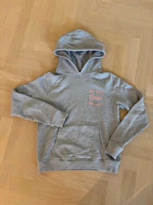 Grå hoodie från Gant med brodyr - Snygg grå hoodie från Gant med rosa broderad text 'New Haven Gant Connecticut' på bröstet. Klassisk känguruficka framtill och mjukt material. Perfekt för en avslappnad och trendig look.