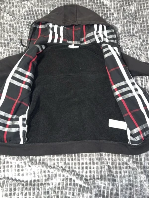 Svart hoodie med rutigt foder - Säljer en svart hoodie med dragkedja och huva. Insidan av huvan och öppningen har ett rutigt foder i svart, vitt, grått och rött som ger en snygg kontrast. Hoodien har dragsko i huvan och ribbade muddar vid ärmar och nederkant. Perfekt för en chill och stilren look.