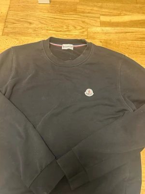 Svart sweatshirt från Moncler - Svart sweatshirt från Moncler med rund halsringning och diskret logotyp på bröstet. Tröjan har långa ärmar och klassisk passform, perfekt för en stilren och avslappnad look.