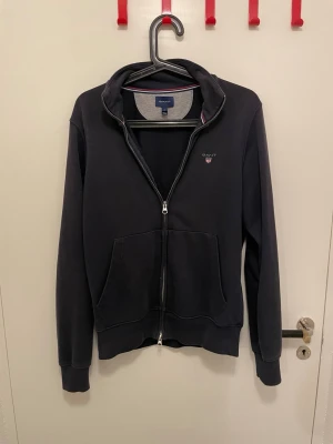 Blå zip tröja från GANT - Snygg blå zip tröja från GANT med dragkedja framtill och två stora fickor. Klassisk logga broderad på bröstet och ribbade muddar vid ärmar och nederkant. Perfekt för en chill och stilren look. 