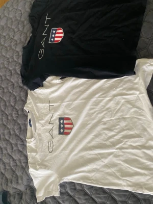 GANT t-shirts med logga och emblem - Två snygga GANT t-shirts, en svart och en vit, båda med GANT-logga och ett emblem med stjärnor och ränder på bröstet. Klassisk passform och korta ärmar, perfekta till jeans eller shorts. Tillverkade i mjuk bomull för skön känsla hela dagen.