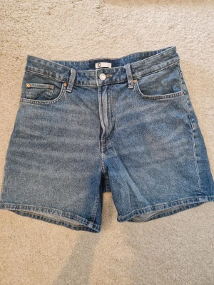 Blå jeansshorts med hög midja - Klassiska blå jeansshorts med hög midja och fem fickor. Shorts i denim med bälteshällor, knappgylf och raka ben. Den sitter som en storlek M men står storlek 30 på den.