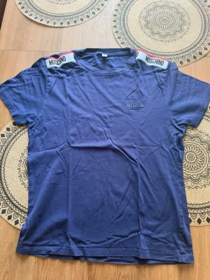Moschino T shirt blå - En mycket fin Moschino tshirt i blå färg. Den är i mycket bra skick. Använd ett fåtal gånger. Nypris på den var 2100kr. 