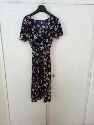 Mörkblå blommig omlottklänning - Supersöt mörkblå omlottklänning med blommigt mönster i vitt och rosa. Klänningen har korta ärmar, v-ringning och knytband i midjan för en snygg passform. Perfekt för sommarens alla tillfällen!