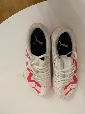 Vita och röda Puma fotbollsskor - Säljer ett par vita fotbollsskor från Puma med röda detaljer och logga. Skorna har snörning, låg profil och en flexibel sula som passar perfekt för inomhusspel. Materialet är syntet med ventilerande meshpartier för extra komfort.
