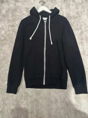 Svart zip hoodie från Pier One - Klassisk svart hoodie från Pier One med dragkedja framtill och vita snören i huvan. Hoodien har två fickor på magen och ribbade muddar vid ärmar och nederkant. Perfekt för en avslappnad och stilren look.
