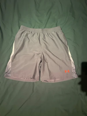 Grå träningsshorts från Under Armour - Snygga grå träningsshorts från Under Armour med elastisk midja och vita detaljer på sidorna. Under Armour-logga i orange framtill och stort tryck längs benet. Perfekta för gymmet eller löpning, tillverkade i lätt och snabbtorkande material.