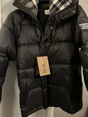 Burberry puffer jacka  - Säljer en svart pufferjacka från Burberry med huva och dragkedja framtill. Jackan har två fickor med dragkedja och en patch med Burberry London England på ärmen. Insidan av huvan har klassiskt rutmönster. Perfekt för kalla vinterdagar.