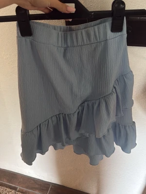 Ljusblå kjol med volangdetaljer - Supersöt ljusblå kjol med volanger och asymmetrisk nederkant. Kjolen har en mjuk struktur och elastisk midja för skön passform. Perfekt att styla med en enkel topp eller stickad tröja.
