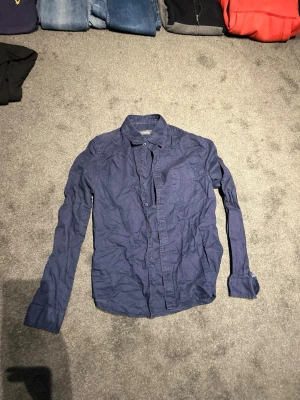 Mörkblå skjorta med lång ärm - Snygg mörkblå skjorta i bomull med klassisk krage och långa ärmar. Skjortan har knappar framtill och en bröstficka. Perfekt till jeans eller chinos för en avslappnad men stilren look.