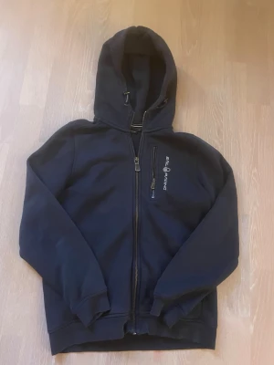 Mörkblå  hoodie från Sail Racing - Mörkblå  hoodie med dragkedja från Sail Racing. Har huva med dragsko, ribbade muddar och en bröstficka med dragkedja. Logga tryckt på bröstet. Perfekt för en avslappnad och sportig stil.