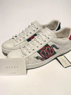 Gucci vita sneakers med orm - Säljer ett par vita Gucci sneakers med den ikoniska gröna och röda randen på sidan och en broderad orm. Baksidan har olika färger, en röd och en grön detalj i ormskinnsmönster. Skorna är låga och har vita snören samt klassisk gummisula.