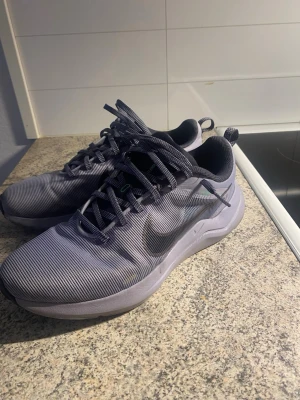 Grå Nike sneakers med svart swoosh - Snygga grå sneakers från Nike med svart swoosh på sidan. Skorna har en sportig design med rund tå, snörning och mesh-liknande material för bra andningsförmåga. Yttersulan är mönstrad för extra grepp och det finns gröna detaljer på plösen och sulan. Den är jätte bra och skön. Har använt den fåtal gånger, nästan som ny. Färgen är unik och speciella, det är typ som ljus lila/grå eller vad nu det är, obs ändå lilla grejen är sista bilden, men det är ingem fara. Några frågor? Skicka pm