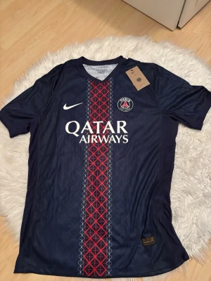 PSG matchtröja Nike mörkblå - Snygg PSG matchtröja i mörkblått med röd och vit detalj i mitten, Qatar Airways-tryck och klubbmärke på bröstet. Tillverkad i lätt och ventilerande material, perfekt för fotboll eller träning. Klassisk Nike-logga på ena sidan.