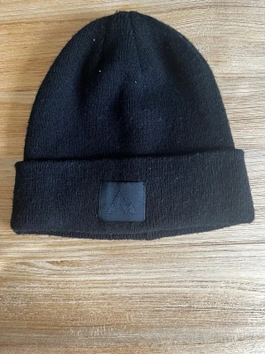 Svart ribbad mössa med patch - Säljer en svart ribbstickad mössa med uppvikt kant och en diskret svart patch framtill med broderad logga. Klassisk modell som passar till det mesta och är enkel att styla. Materialet känns mjukt och stretchigt, perfekt för kyliga dagar.