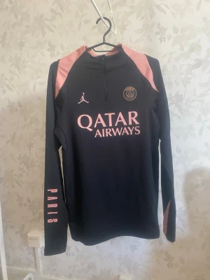 Jordan svart träningströja, storlek S - Jordan svart träningströja, storlek S. Snygg långärmad träningströja från PSG i svart med rosa detaljer på axlar och ärmar. Tröjan har halv dragkedja framtill, tryck med Qatar Airways och Paris Saint-Germain-logga samt texten PARIS på ena ärmen. Perfekt för fotbollsträning eller chill.