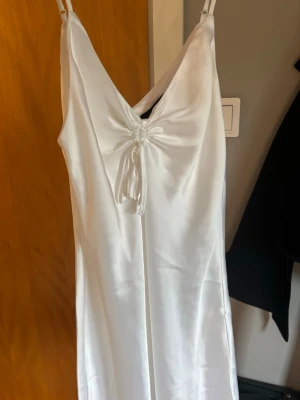 Vit långklänning i satin från BikBok - Elegant vit långklänning från BikBok i glansigt satinmaterial. Klänningen har smala axelband, v-ringning och en snygg detalj med knyt framtill. Perfekt för sommarens speciella tillfällen.