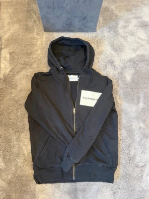 Svart hoodie med dragkedja Calvin Klein - Snygg svart hoodie från Calvin Klein med dragkedja framtill och stor vit logga på bröstet. Sizen är M och skicket är 9/10
