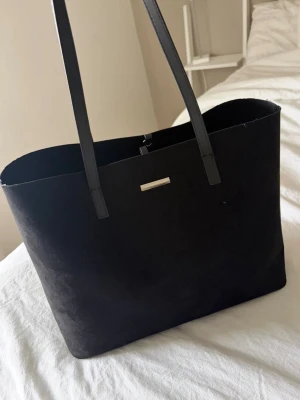 Svart shoppingväska i mockaimitation - Svart väska perfekt till skolan. Väskan har en mocka design och silver detaljer. Måtten kan man se på bilderna, det får enkelt plats en skoldator, böcker och block. Långa band 
