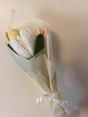 Virkat blomsterbukett i pastell - Söt bukett med virkade tulpaner i pastellfärger – vitt, gult och ljusrosa, med gröna blad. Blommorna är gjorda i garn och inslagna i silkespapper med ett vitt snöre. Perfekt som dekoration eller present till någon du gillar.