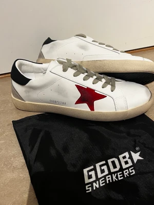 Vita GGDB sneakers med röd stjärna - Snygga vita sneakers från Golden Goose med en ikonisk röd stjärna på sidan, beige sula och grå snören. Skorna har svart detalj på hälen och är tillverkade i läder. Perfekta för dig som gillar en clean men ändå unik look.