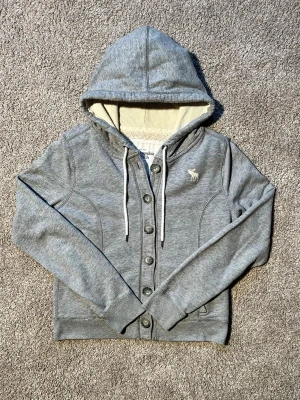Grå hoodie med knappar och huva - Snygg grå hoodie från Abercrombie & Fitch med knappar framtill, vit dragsko i huvan och mjukt, ljust innerfoder. Klassisk logga broderad på bröstet och ribbade muddar vid ärmar och nederkant. Perfekt för en avslappnad och trendig look. Storlek L enligt etiketten men sitter som en S-M