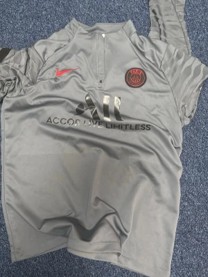 Grå PSG träningströja med halv dragkedja - Snygg grå träningströja från PSG med halv dragkedja framtill. Tröjan har svarta och röda detaljer, PSG-logga på bröstet och Nike-logga i rött. Tillverkad i ett lätt och funktionellt material, perfekt för träning eller sport.