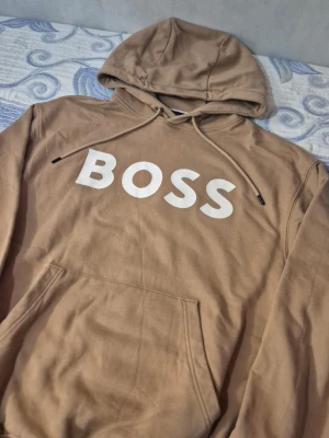 BOSS hoodie - Snygg beige hoodie från BOSS med stor vit logga på bröstet. Hoodien har huva med dragsko och en klassisk magficka. Tillverkad i mjukt bomullsmaterial som känns skönt mot huden. Perfekt för en avslappnad och trendig look.