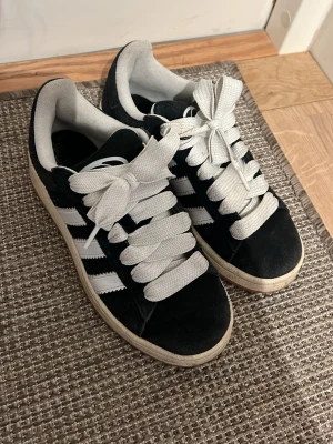 Adidas campus - Snygga svarta sneakers med breda vita skosnören och klassiska vita ränder på sidorna. Skorna har låg profil och en vit sula som ger en retro känsla. Ovandelen är i mockaliknande material och insidan är vadderad för extra komfort. Något slitna o lite fläckiga som syns på bilderna