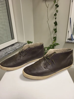 Fred Perry Chukka Kängor Bruna Storlek 43 - Säljer ett par bruna loafers i läder med vita sulor och gröna snören. Skorna har en enkel och stilren design med låg profil och mjukt innerfoder. Perfekta för dig som gillar klassisk men avslappnad stil.