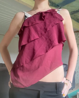 Vinröd volang topp med spetsband - Säljer en vinröd topp med flera volanger och spetsdetaljer på axelbanden. Toppen är ärmlös och har en asymmetrisk nederkant som ger en unik look. Perfekt för dig som vill sticka ut lite extra.