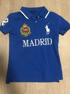 Blå pikétröja från Ralph Lauren - Snygg blå pikétröja från Ralph Lauren med vit krage, broderad logga och emblem på bröstet samt texten 'MADRID'. Stor vit siffra 2 på ryggen och ärmen. Klassisk piké med korta ärmar och knappar vid halsen.
