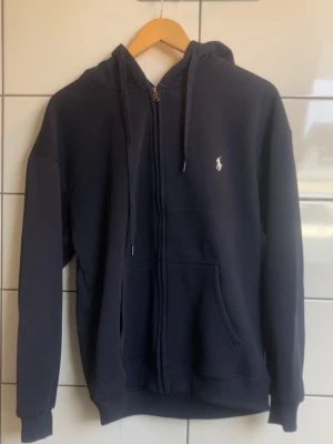 Polo Ralph Lauren Zip Hoodie i mörkblå färg. - Storlek M.  Endast provad, aldrig använd. Säljs utan prislapp och kvitto. Hoodien är i mycket fint skick utan hål, fläckar eller andra anmärkningar.  Klassisk modell med broderad Polo Pony-logga på bröstet, dragkedja framtill, huva och framfickor.  Passar perfekt till både vardags- och mer uppklädda outfits.  Skicka gärna meddelande vid frågor eller om du vill ha fler bilder.  Öppen för seriösa bud.