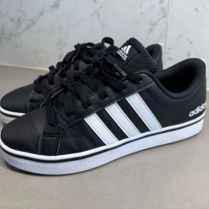 Svarta Adidas sneakers med vita ränder - Klassiska svarta Adidas sneakers med de ikoniska tre vita ränderna på sidan. Skorna har svart ovandel i syntetmaterial, svart snörning och vit sula. Perfekta för en clean och sportig look. Adidas-logga på plösen och hälen.