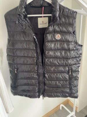 Svart dunväst från Moncler - Snygg svart dunväst från Moncler med glansig finish och klassisk quiltad design. Västen har två dragkedjeförsedda fickor framtill, hög krage och Moncler-logga på bröstet. Perfekt för lager-på-lager och streetstyle. Fickan på insidan är sönder