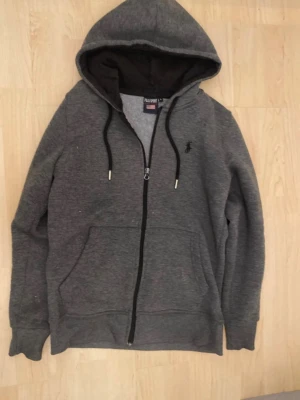 Mörkgrå polo hoodie med huva strl 160 - Mysig mörkgrå hoodie med dragkedja framtill och stor känguruficka. Justerbar huva med svarta snören och ribbade muddar vid ärmar och nederkant. Liten broderad logga på bröstet. Perfekt för chill dagar eller till vardagslooken.