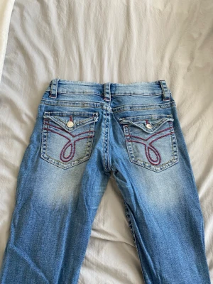 Blå slitna jeans med broderade fickor - Säljer ett par ljusblå jeans med slitningar på knäna och snygga broderade detaljer i rött på bakfickorna. Jeansen har bootcut-modell och klassisk femficksdesign. Perfekta för en avslappnad och trendig look.