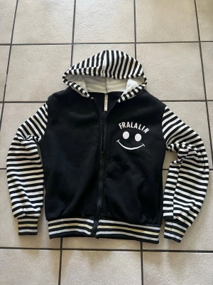 Svart hoodie med ränder och smiley storlek M/L - Säljer en svart hoodie med dragkedja och huva. Ärmar och huva har svartvita ränder, och på bröstet samt ryggen står det 'FRALALIN' med en stor vit smiley. Ribbstickade muddar och nederkant med randigt mönster. Perfekt för dig som gillar en lekfull och unik stil.
