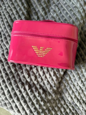 Rosa/röd  necessär Emporio Armani - Snygg rosa necessär från Emporio Armani i glansigt material med guldfärgade detaljer och dragkedja. Perfekt för smink eller småprylar. Logga framtill och två guldiga dragkedjor baktill ger en lyxig känsla.