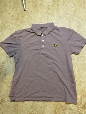Lyle & Scott pikétröja med krage - Snygg lila pikétröja med klassisk krage och tre knappar framtill. Tröjan har korta ärmar och en gul broderad logga på bröstet. Perfekt för en avslappnad och stilren look.