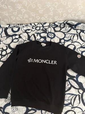 Svart Moncler sweater med logga - Säljer en svart Moncler sweater med stor vit logga på bröstet och klassisk patch på ärmen. Hoodien har rund halsringning och långa ärmar. Perfekt för dig som vill ha en clean och stilren look.