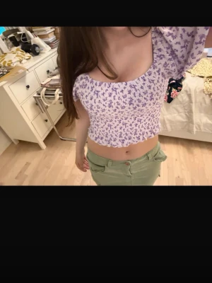 Ljuslila blommig croppad topp - Supersöt ljuslila croppad topp med blommigt mönster och puffärmar. Toppen har smockad midja och volangkant nedtill, vilket ger en feminin och trendig look. Perfekt för vår och sommar!