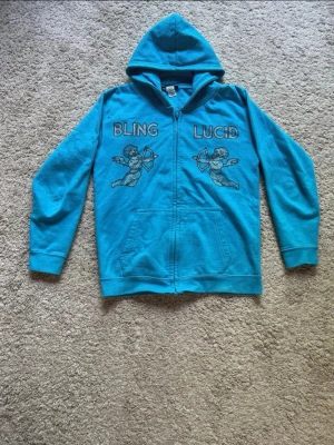 Turkos/ljusblå blinglucid Hoodie 140 - Säljer en blå rinestone Hoodie från bling Lucid i storlek 140. Det är en cool och unik design som sticker ut och färgen gör att stenarna blänger i ljuset. Den är knappt använd och håller hög kvalitet, men var försiktig när du tvättar den