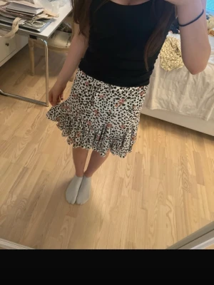 Kort volangkjol med leopard- och blommönster - Säljer en kort kjol med volangkant och elastisk midja. Kjolen har ett unikt leopardmönster i svart och vitt, kombinerat med små blommor i rosa och orange toner. Perfekt för dig som vill sticka ut med en trendig och lekfull stil.