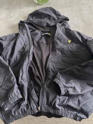 lyle n scott vind jacka  - Snygg svart vindjacka med huva och dragkedja framtill. Jackan har resår vid ärmslut och nederkant samt ett gult broderat märke på bröstet. Tillverkad i lätt och vindtätt material, perfekt för blåsiga dagar.
